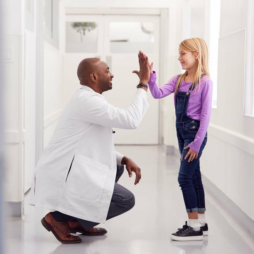 Male Pediatric Doctor Giving Young Girl Patient High Five In Hospital Corridor